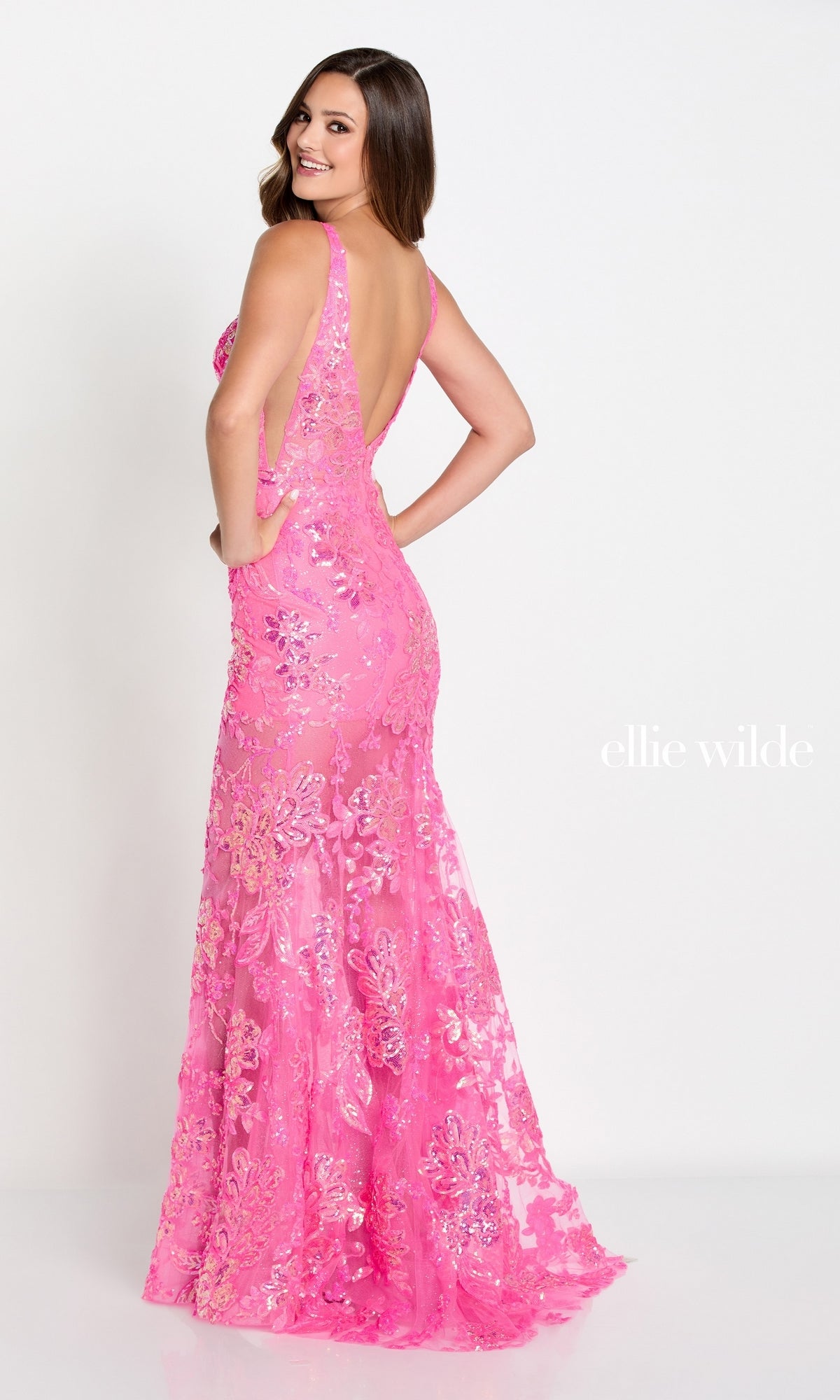 A woman models the Ellie Wilde Sheer-Skirt Long Prom Dress EW34040—a floor-length, sleeveless, backless pink sequin mermaid gown with floral patterns—smiling slightly away against a plain white background. The text reads